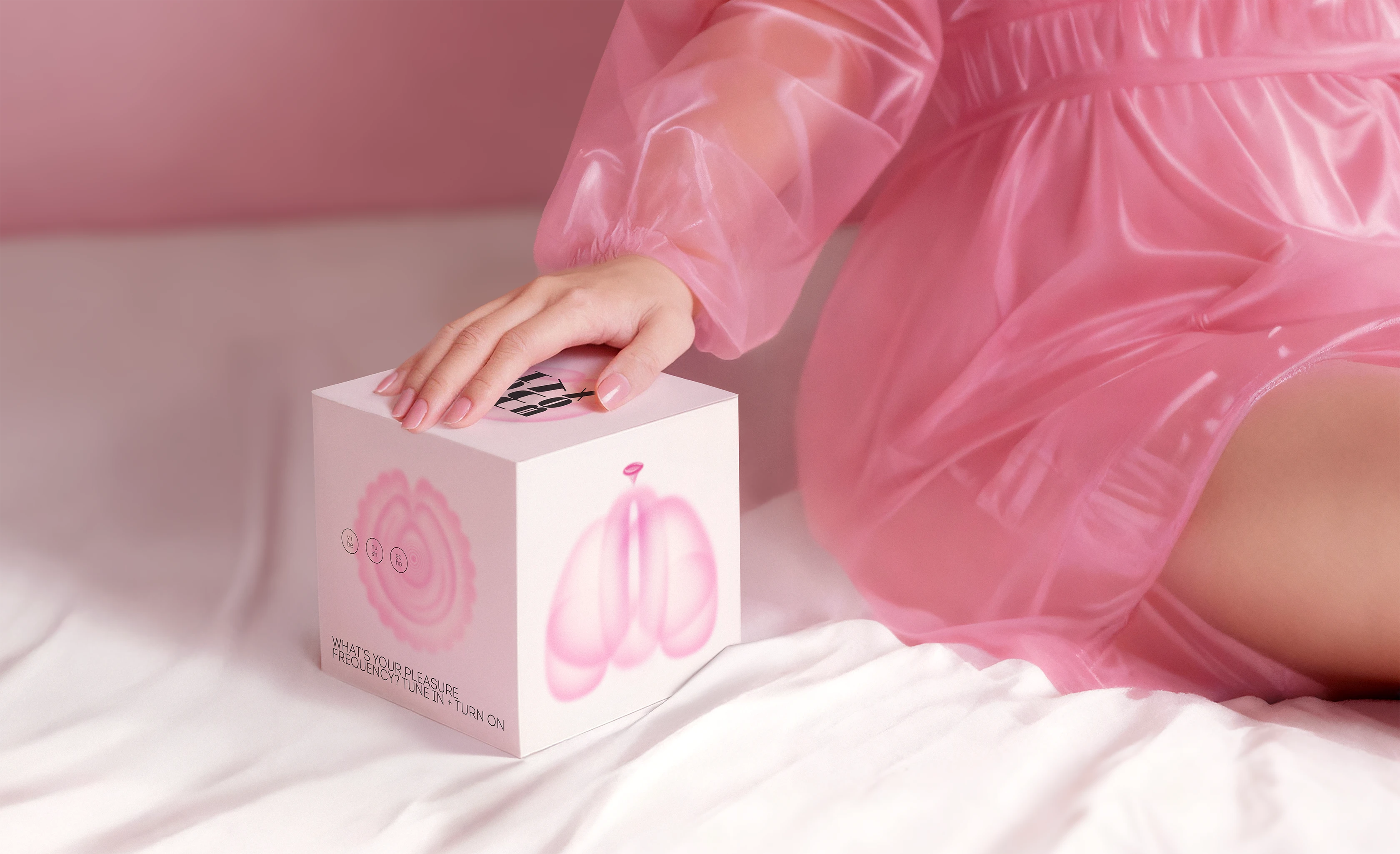 Close-up of a hand resting on the Oh My Box! package while wearing a pink translucent dress, seated on a soft bed.