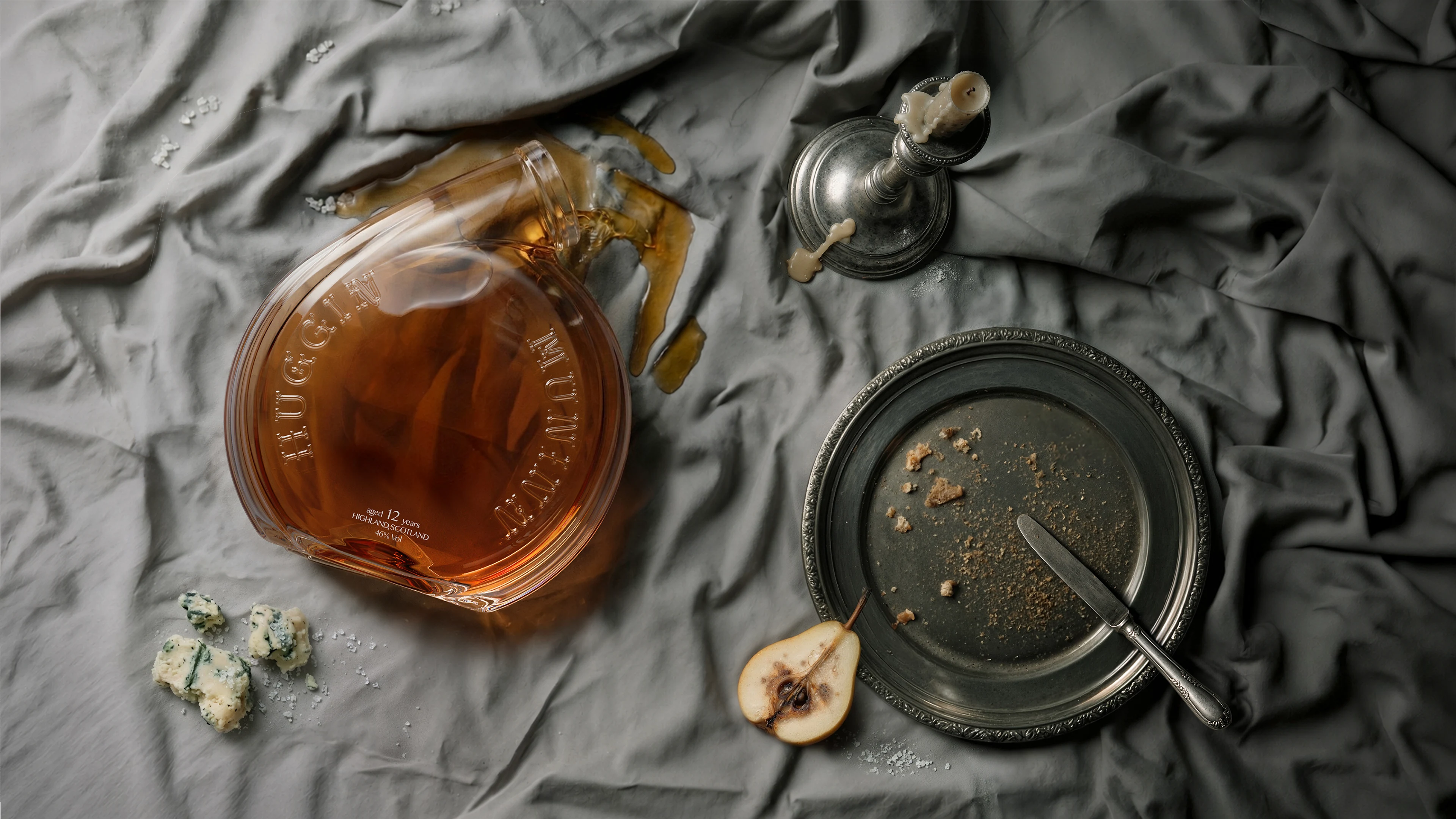 Overhead flat-lay of award-winning creative packaging design for Huginn & Muninn whisky by DEEEZ Design & Creativity Ltd, showcasing an open embossed glass bottle spilling amber spirits across draped linen in a Vanitas still-life scene.