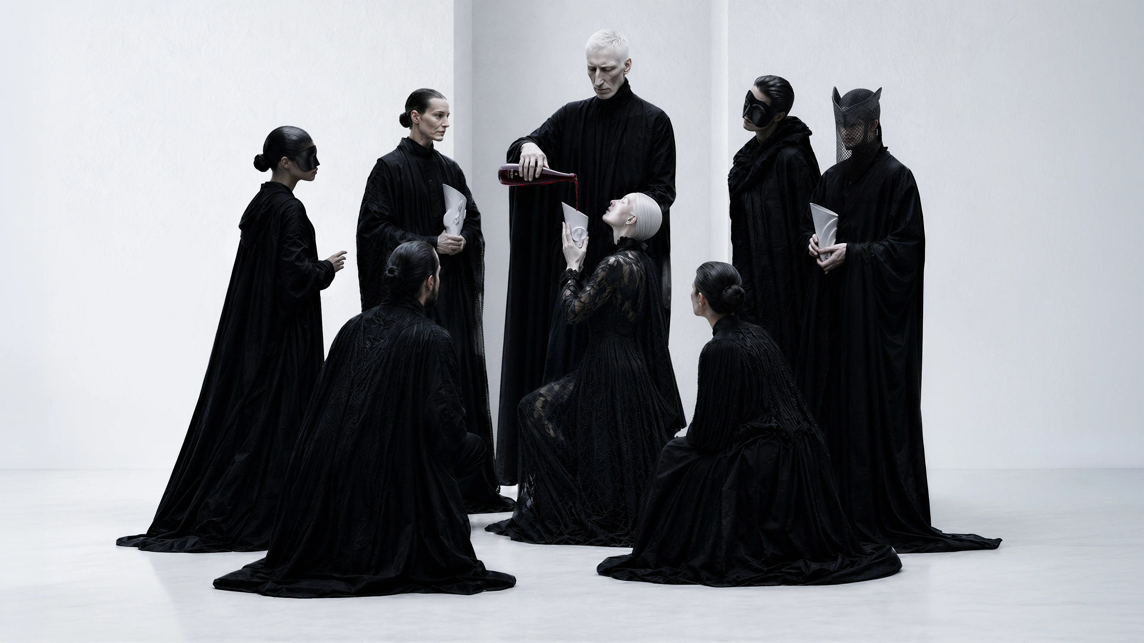 People in black robes gather as red liquid is poured from a RHEYN bottle into a white sculptural drinking vessel.