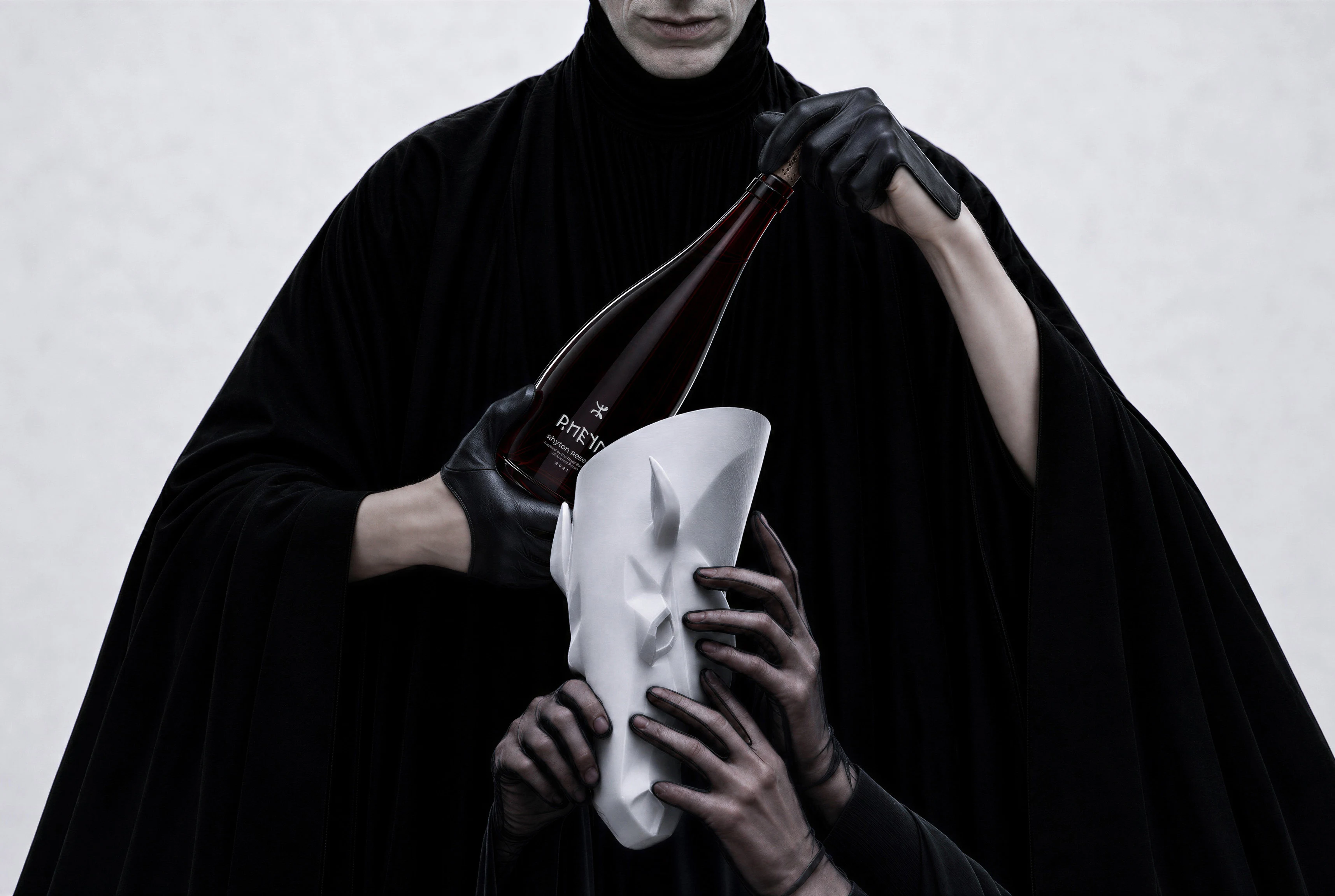 Black-gloved hands present a deep red RHEYN bottle beside its white sculptural animal-head cover.